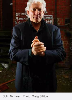 Colin McLaren, silver-haired and dressed in dark clothing, standing with his hands clasped in front of a graffitied brick wall. Photo: Craig Sillitoe.