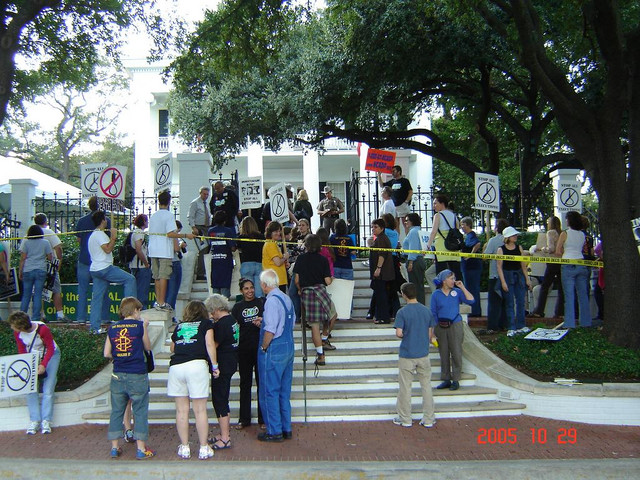 Anti-death penalty marchers gather at the front gate of the Texas Governor's Mansion in October 2005, many holding "Stop All Executions" placards showing a crossed-out noose, with a state trooper at the gate and yellow tape across the steps.