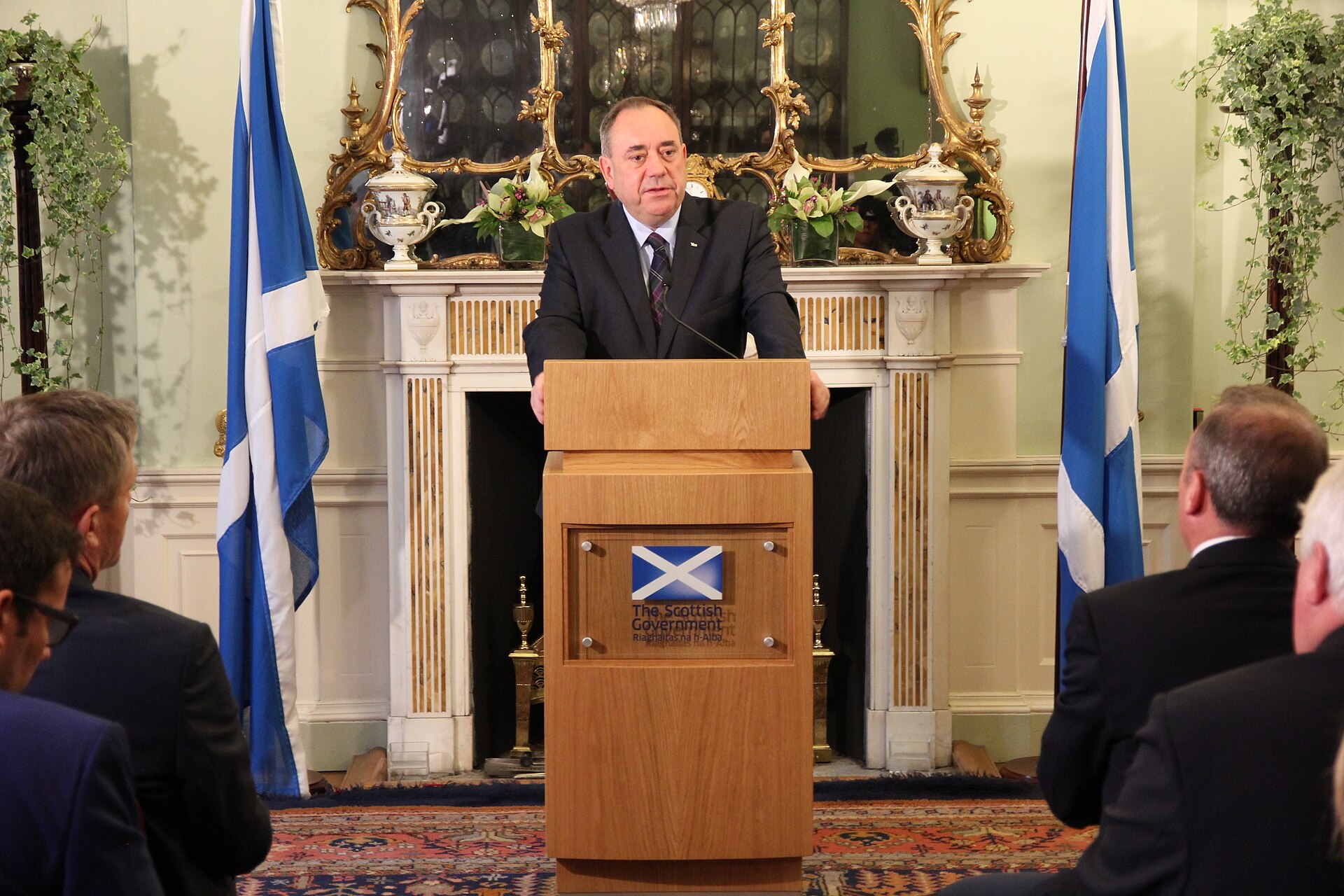 Alex Salmond, in a dark suit and tie, speaking at a Scottish Government lectern in a formal room with a gilt mirror, fireplace and two Saltire flags, as an audience watches.