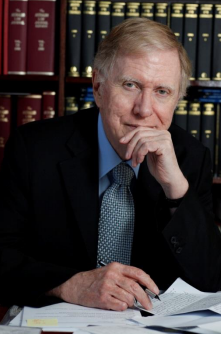 The Hon Michael Kirby AC CMG seated at a desk of papers, chin resting on one hand, in front of shelves of law reports.