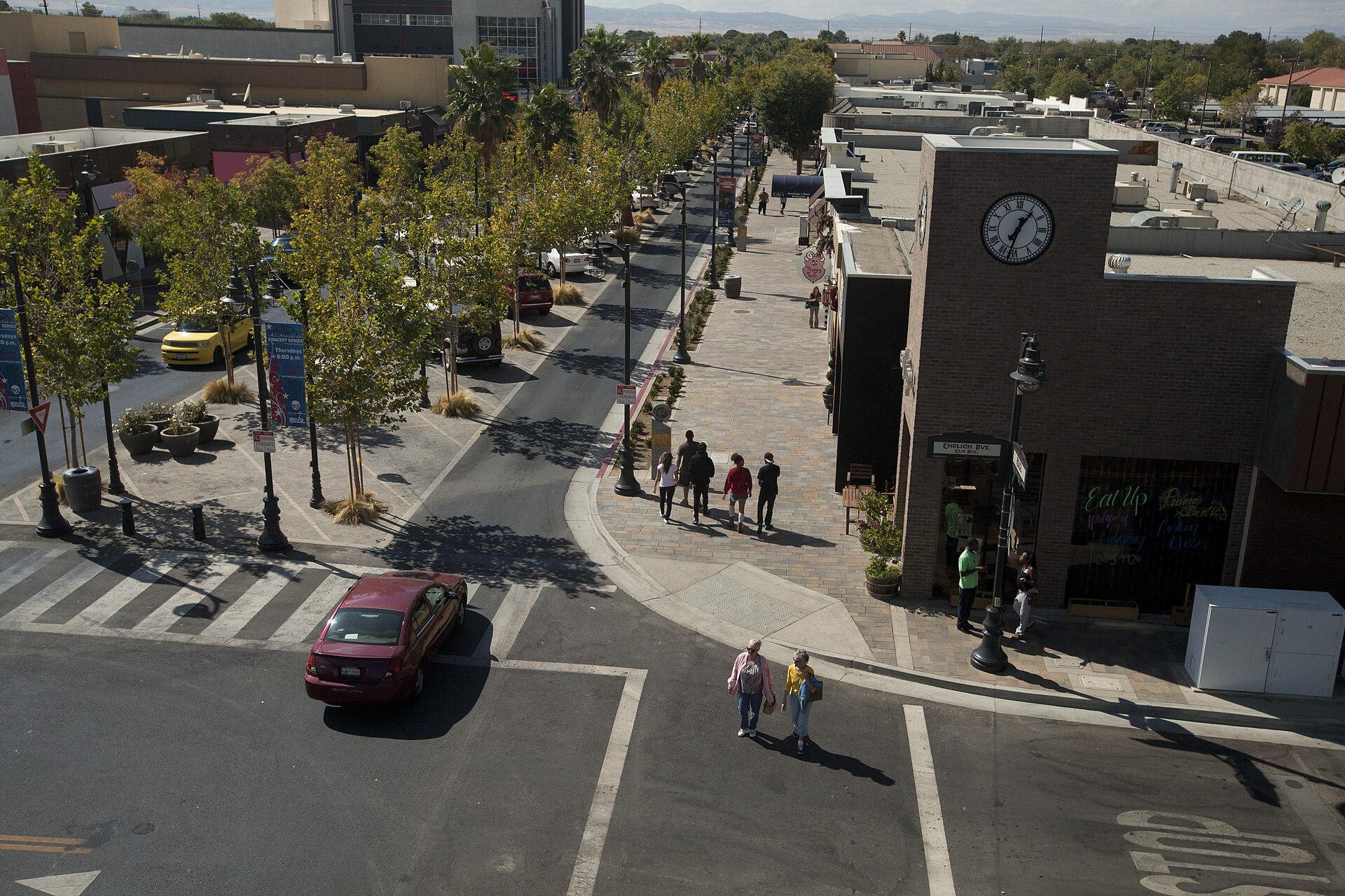 Looking down on Lancaster Boulevard in Lancaster, California: a tree-lined street with a brick clock tower on the corner, pedestrians crossing and walking the pavement, and desert mountains on the horizon.