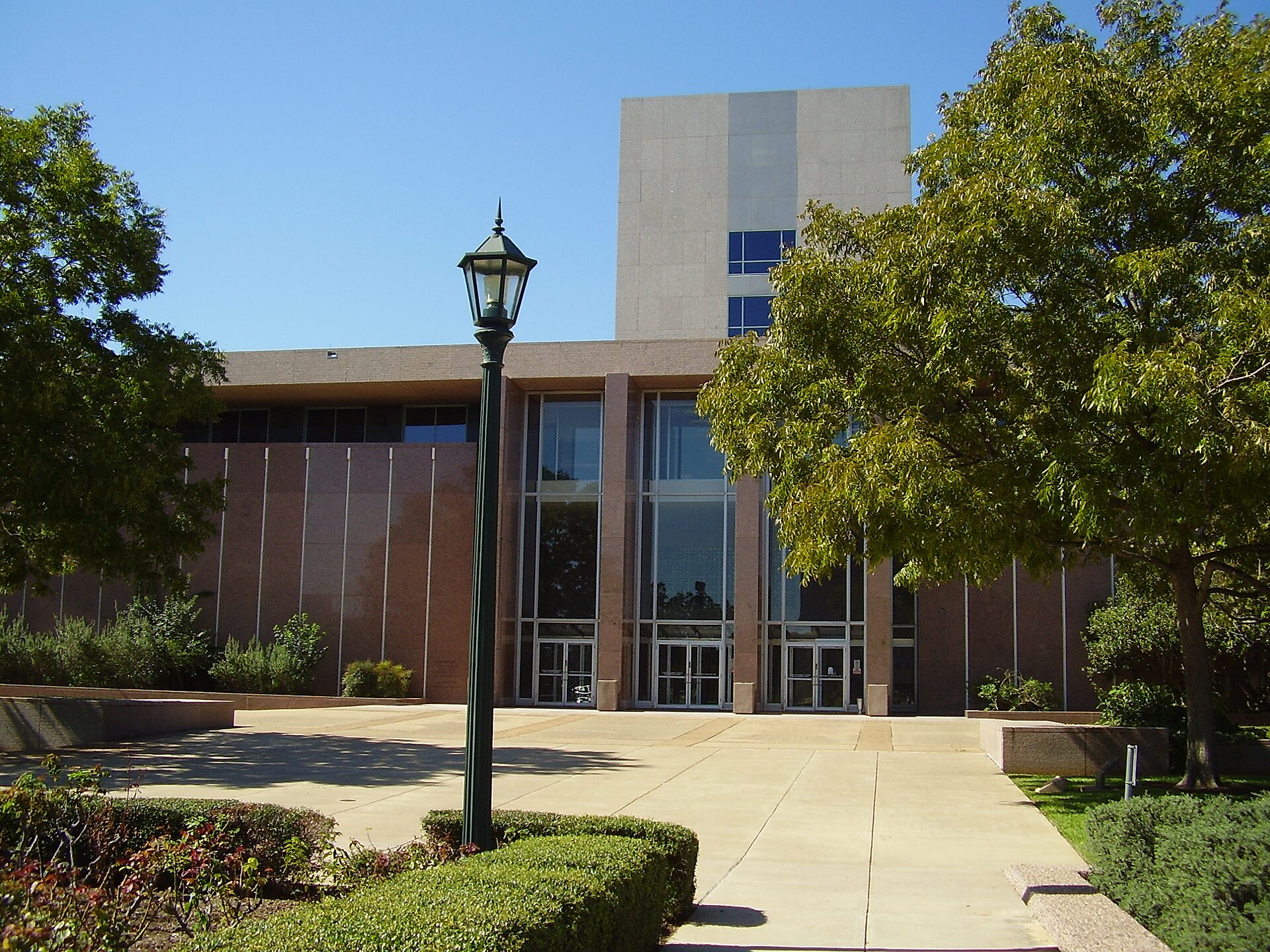 A low pink granite building with a tall glass entrance and a pale stone tower behind it, framed by trees and a lamp post, at the Texas Supreme Court Building in Austin.