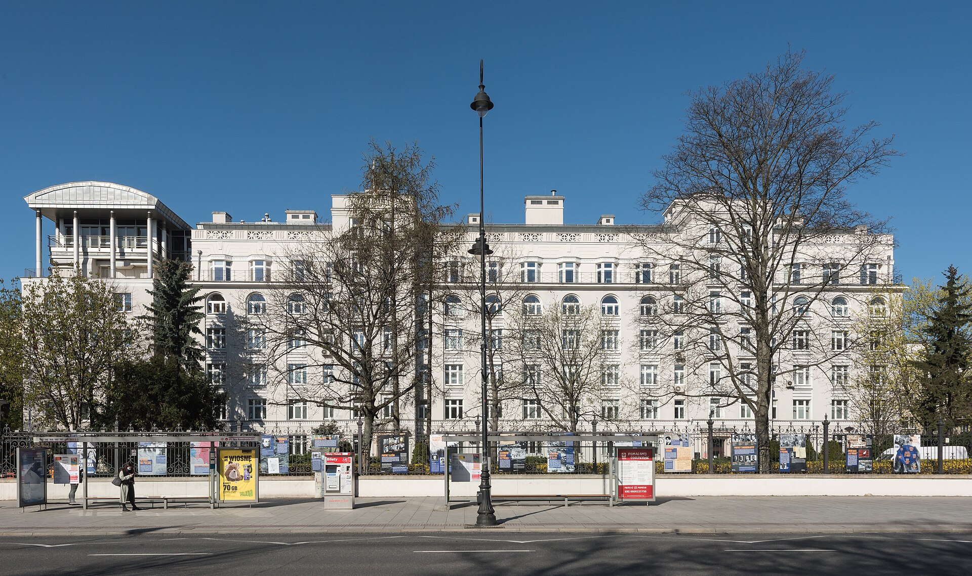 The long white facade of Poland's Ministry of Justice on Aleje Ujazdowskie in Warsaw, seen across the street behind railings, bare trees and a tram stop, under a deep blue sky.