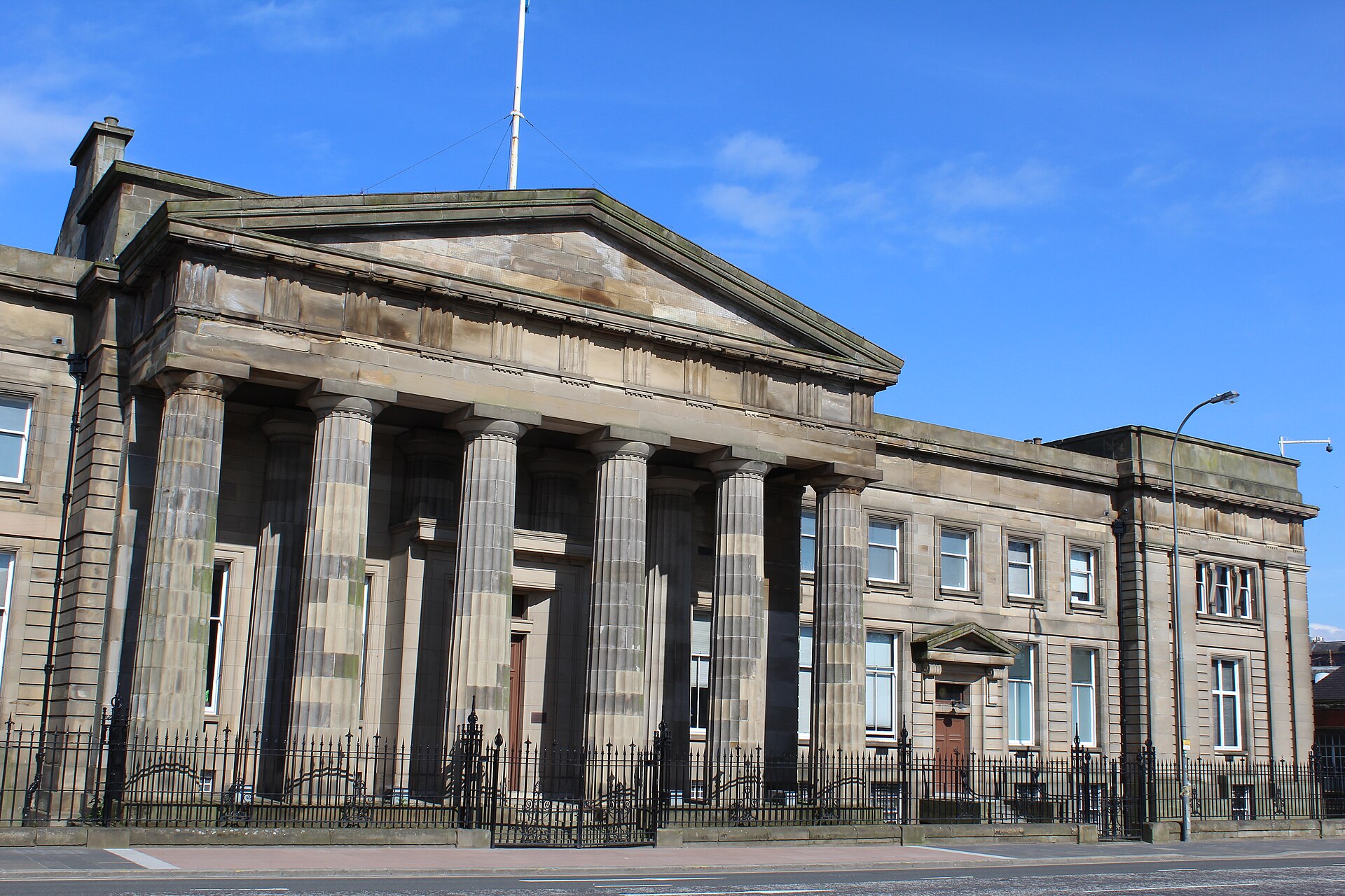 The sandstone front of the Justiciary Courthouse on Saltmarket, Glasgow: a pedimented portico of six fluted Doric columns behind black iron railings, under a blue sky.
