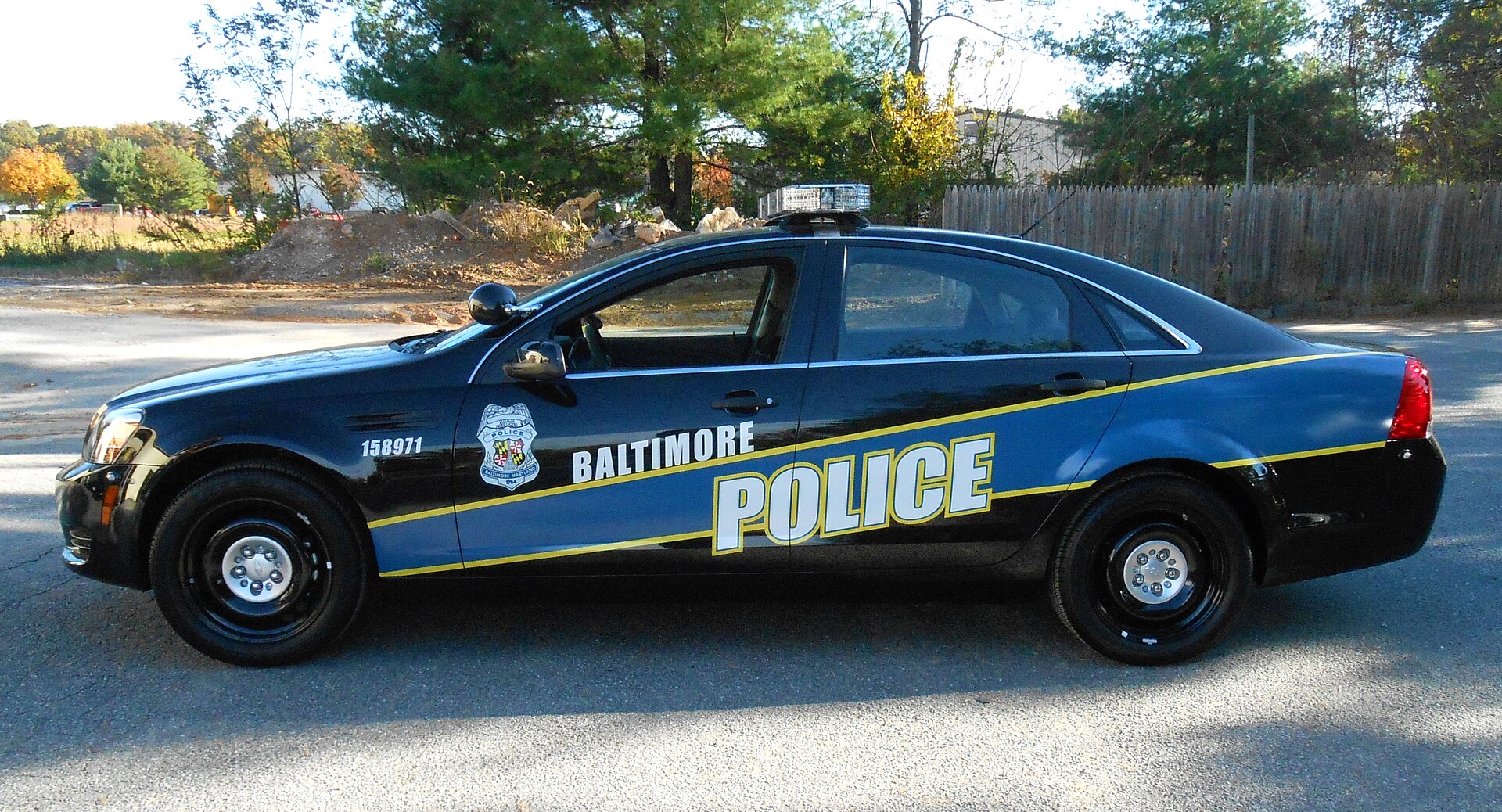 A Baltimore Police Department Chevrolet Caprice patrol car, painted black and blue with a gold stripe, 'BALTIMORE POLICE' lettering and the department badge on its doors, parked on a gravel lot.