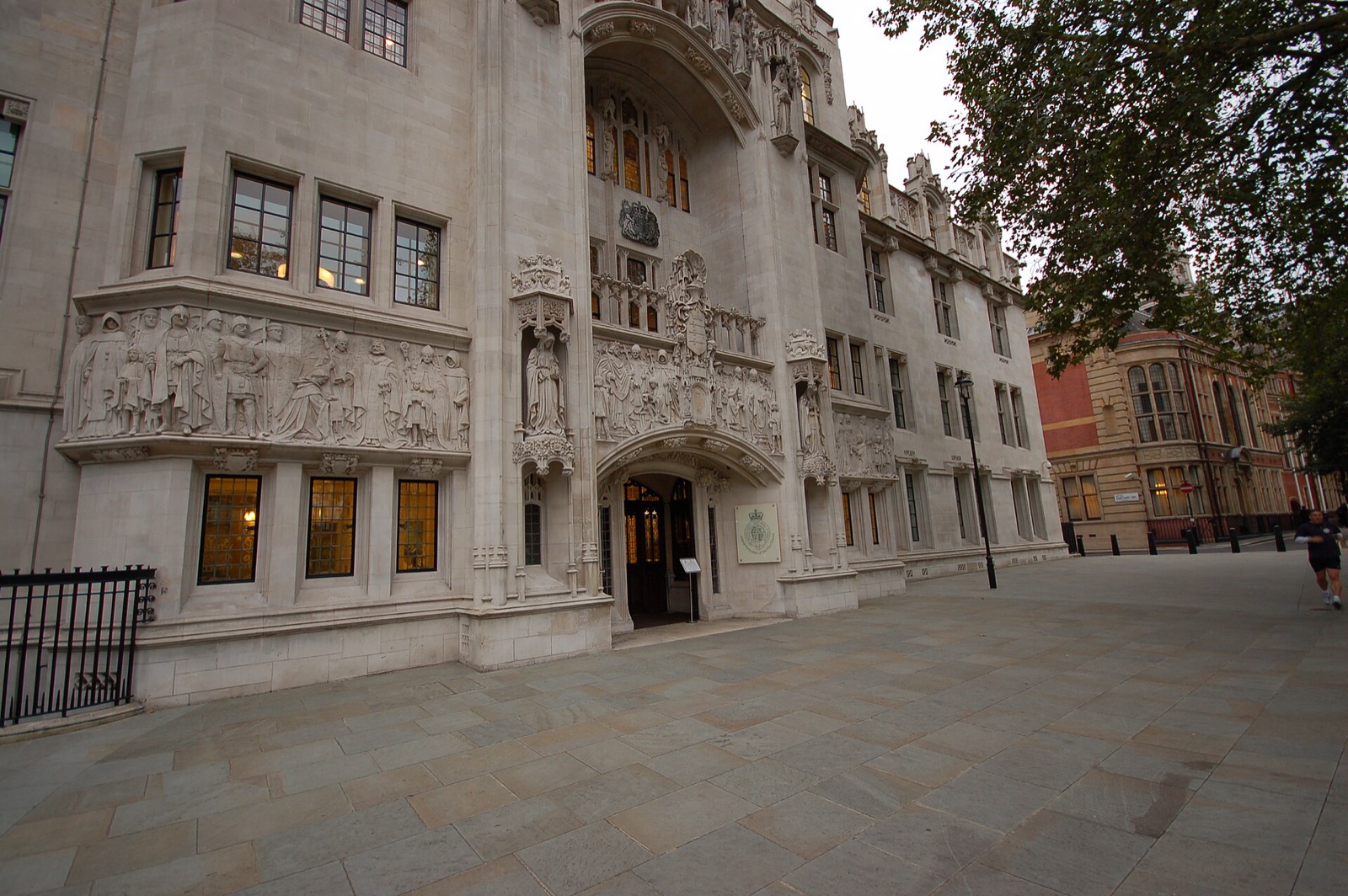 The carved stone entrance of the UK Supreme Court in the Middlesex Guildhall on Parliament Square, London, with a frieze of sculpted figures and statues framing a lit doorway, beside a paved square at dusk.