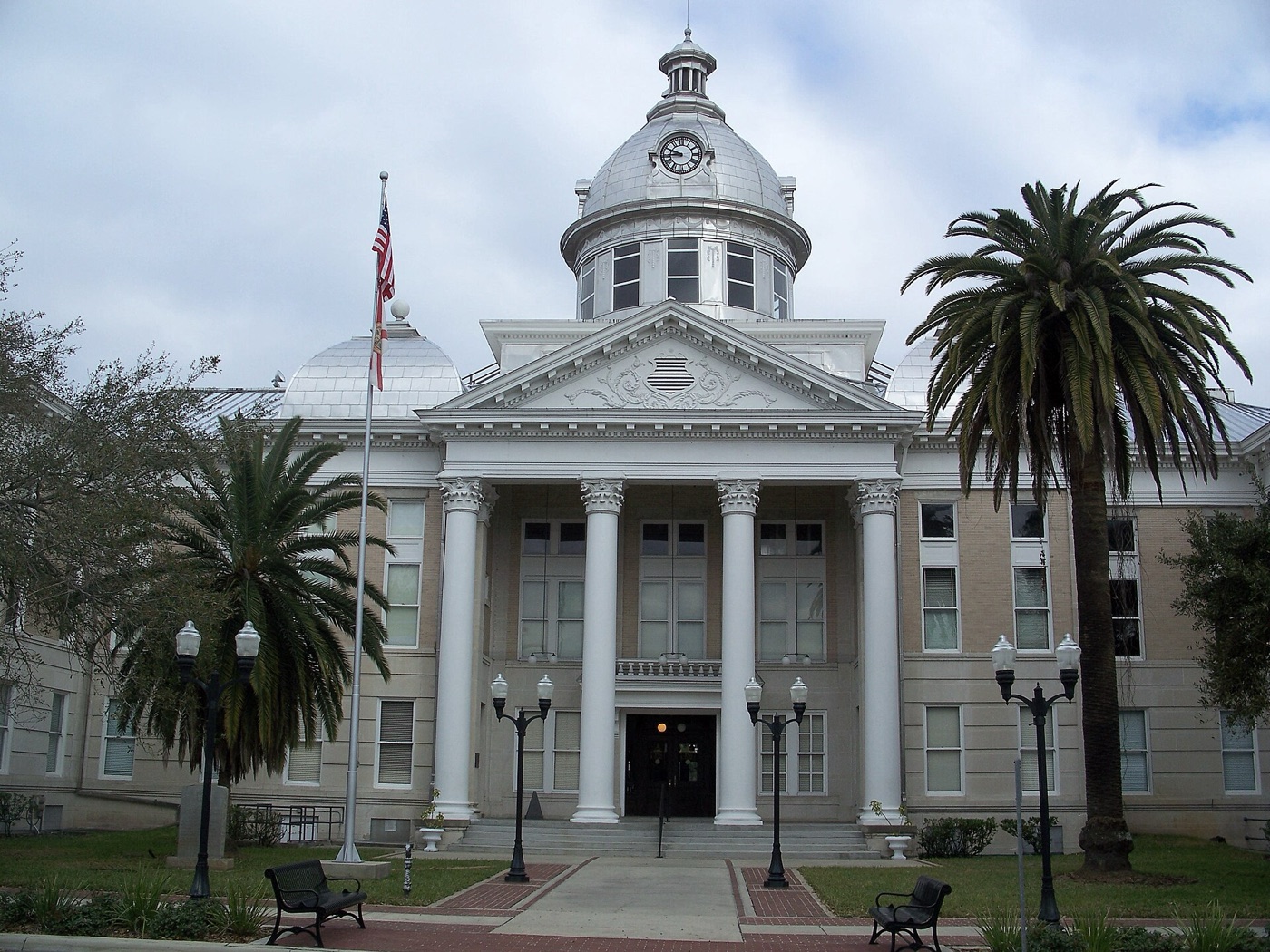 The Old Polk County Courthouse in Bartow, Florida, seen head-on - a cream neoclassical building with a tall silver clock dome, a pedimented portico on Corinthian columns, palms to either side and a US flag on a pole in front.