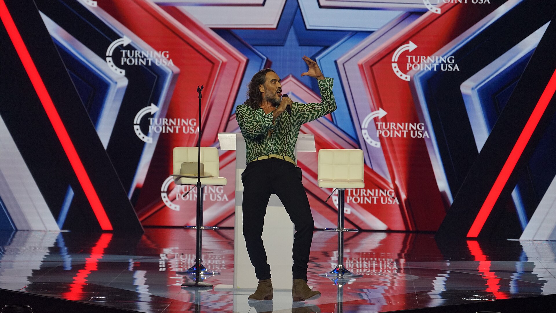 Russell Brand in a green-and-white patterned shirt and dark trousers speaks into a microphone on a stage, one hand raised, between two white bar stools, against a red, white and blue backdrop.