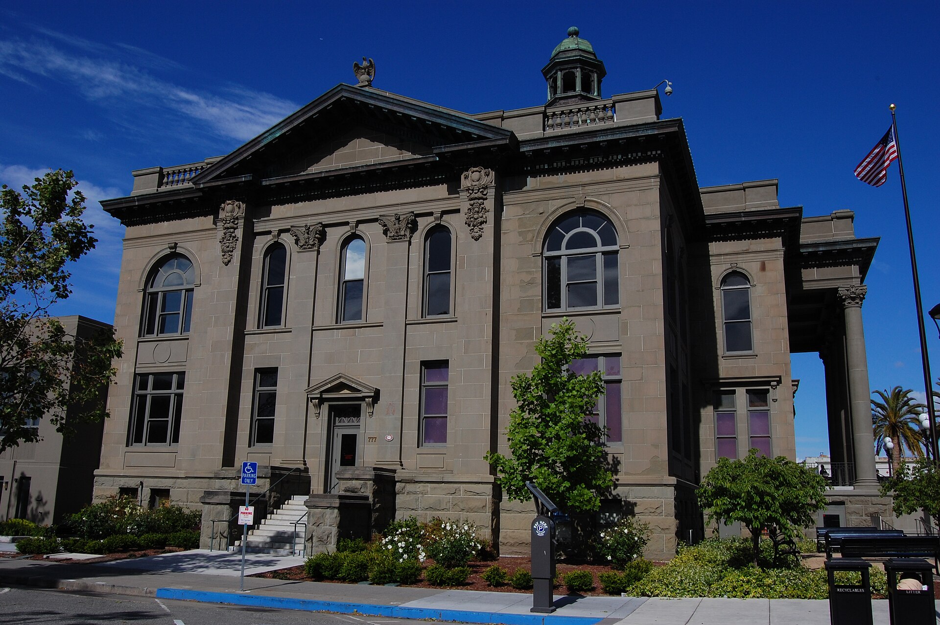 The grey stone front of the historic San Mateo County Courthouse in Redwood City, with arched windows, a pediment topped by an eagle, a green cupola and an American flag, under a blue sky.