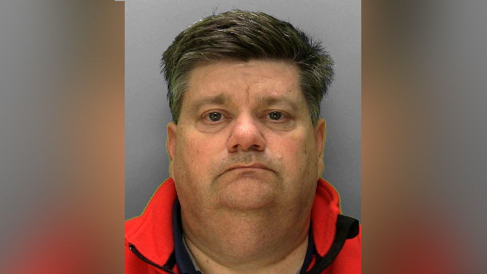 Police custody photograph of Carl Beech, a heavy-set man with short greying dark hair, wearing a red fleece, against a grey background.