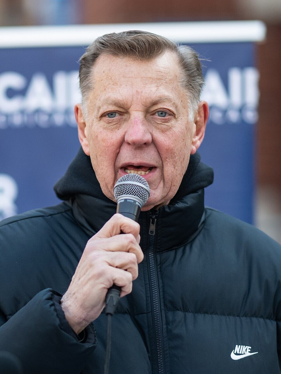 The Rev. Michael Pfleger speaking into a handheld microphone at an outdoor event, wearing a black padded jacket, with a CAIR banner behind him.