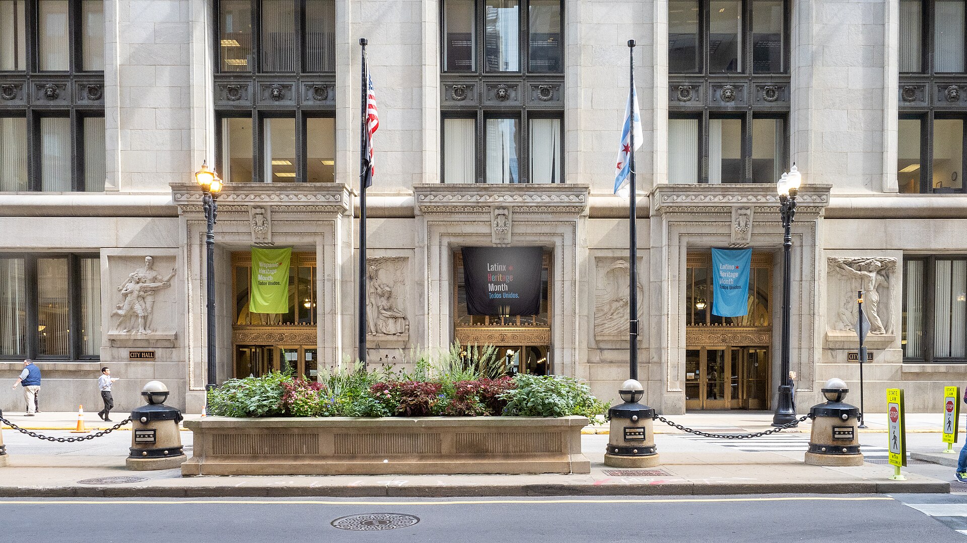 The LaSalle Street entrance to Chicago City Hall, a grey limestone building with carved CITY HALL plaques flanking three bronze doorways, the American and Chicago flags flying outside.