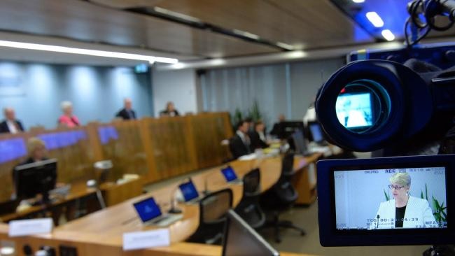 A television camera in the foreground records a witness giving evidence at a royal commission hearing table.