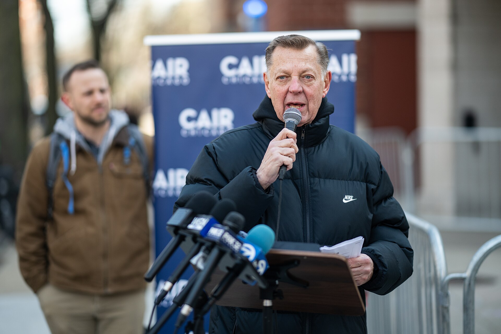 Father Michael Pfleger speaking into a microphone at an outdoor press conference, in front of a CAIR Chicago banner and a bank of television news microphones