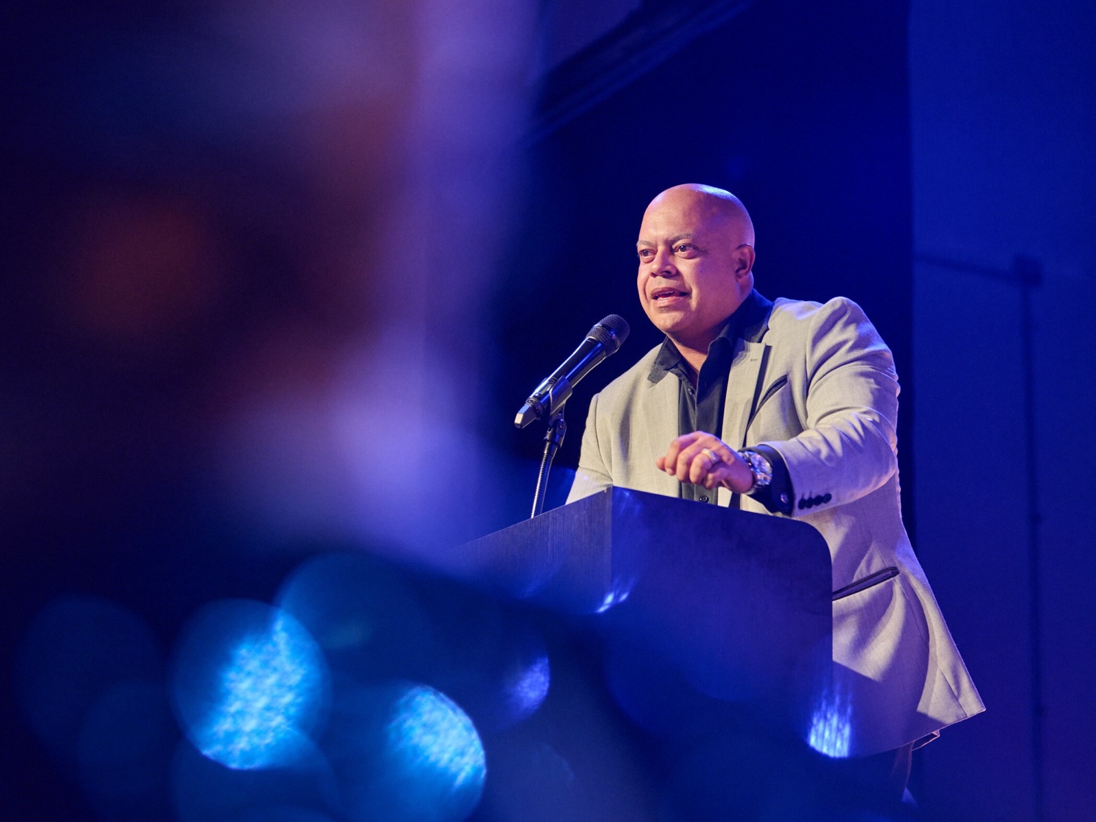 Jon-Adrian 'JJ' Velazquez, a bald man in a light grey jacket over a dark shirt, speaking at a lectern with a microphone, lit in blue on stage.