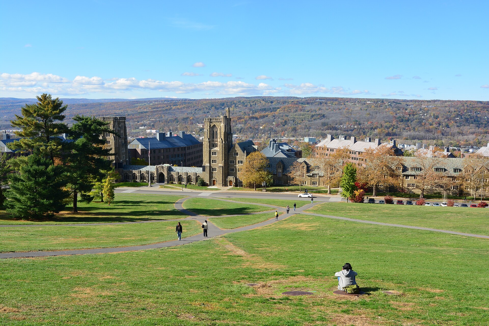 Looking down the grassy Libe Slope at Cornell University towards West Campus, with Gothic stone residence halls and a tower among autumn trees, hills beyond under a blue sky, and a few students on the paths.