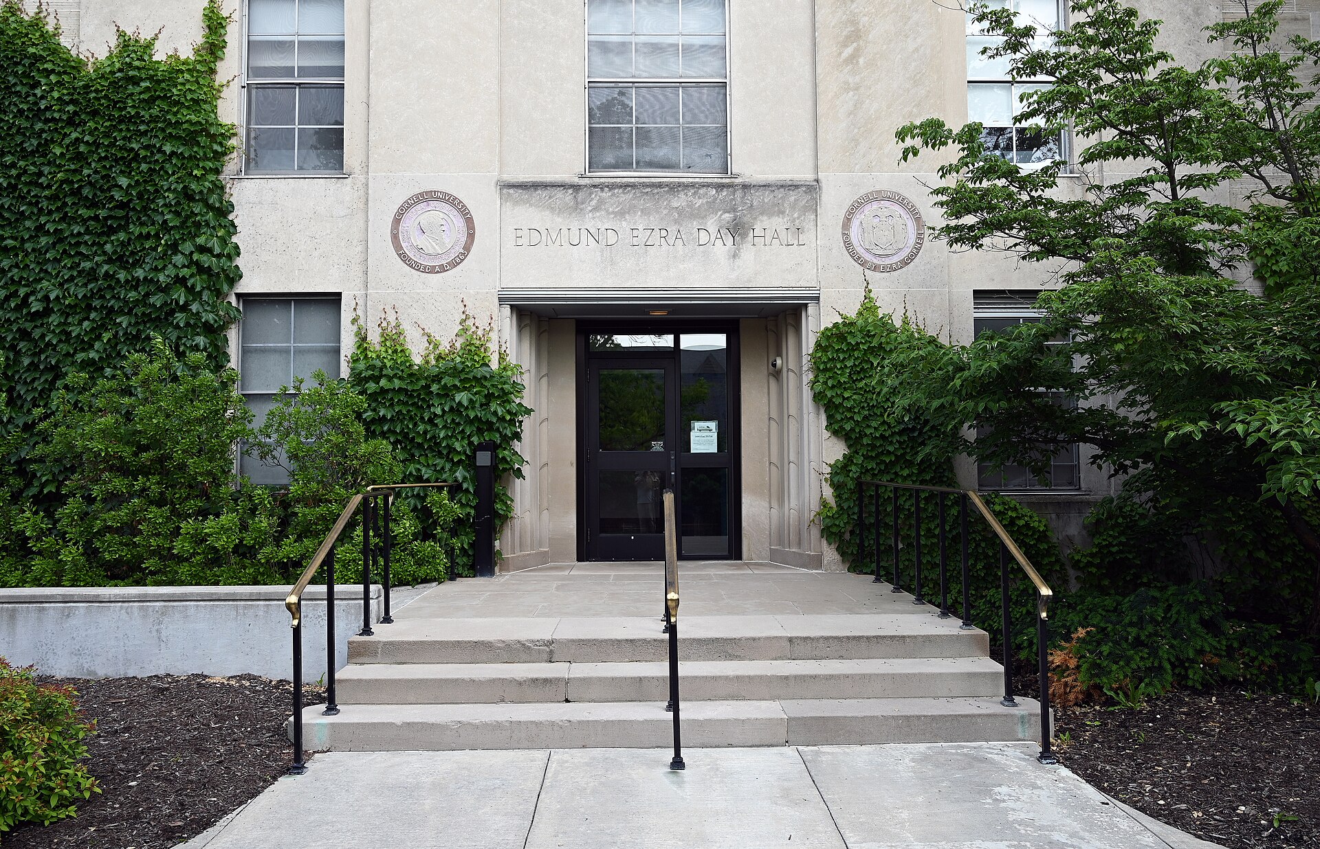 The main entrance of Day Hall at Cornell University, with EDMUND EZRA DAY HALL carved above the doorway between two university seals, ivy on the pale stone walls and steps with brass handrails leading up.