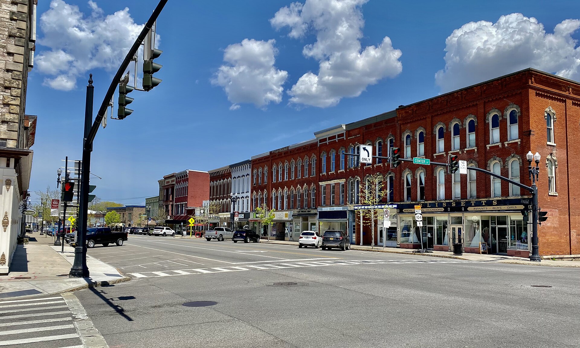 Main Street in Medina, New York, lined with two- and three-storey red-brick Italianate shopfronts under a blue sky with white clouds, seen from the Center Street crossing.