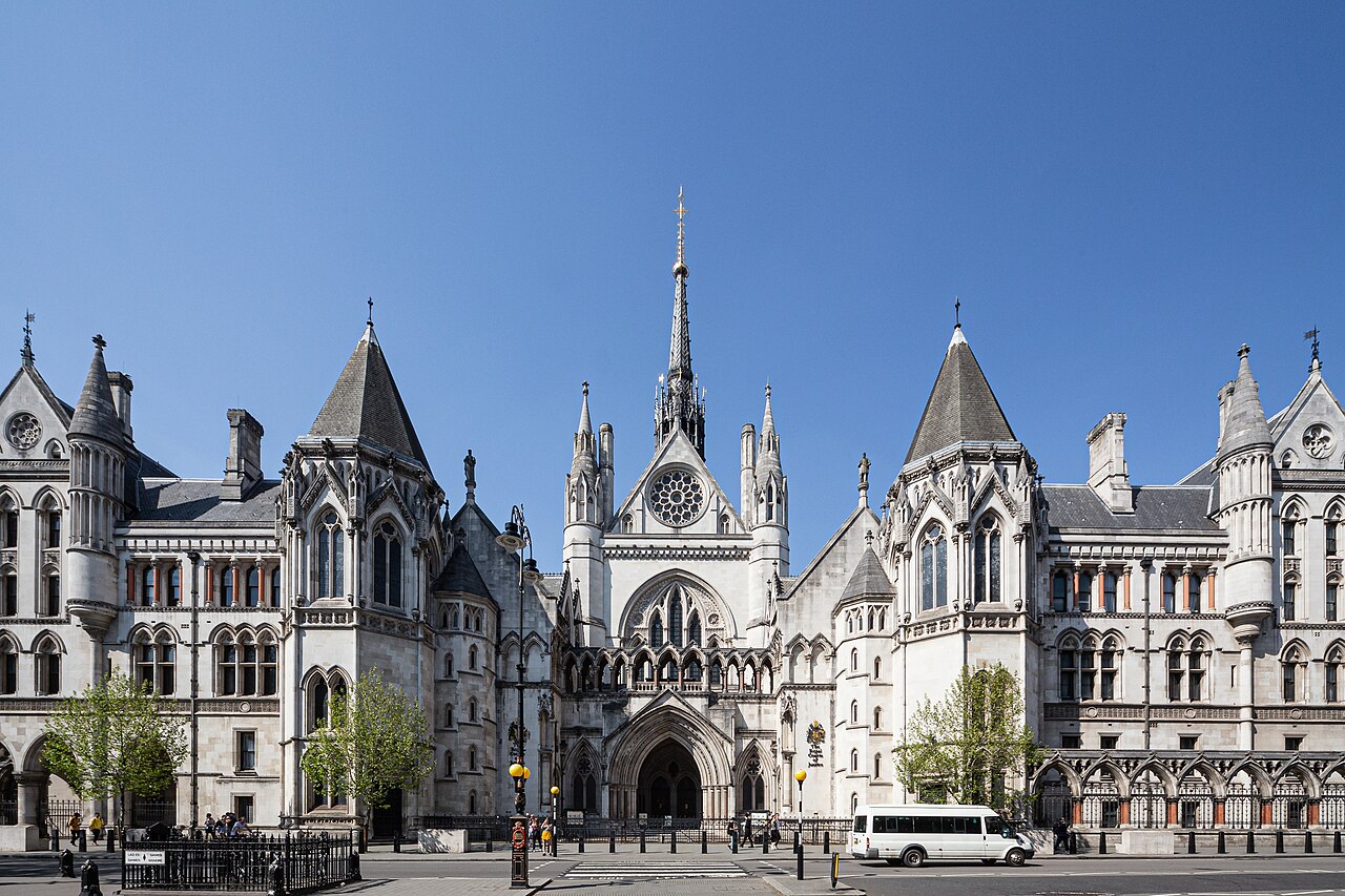 The main gate of the Royal Courts of Justice on the Strand in London, a white Victorian Gothic stone building with a central spire, pointed turrets and arched windows, under a clear blue sky.
