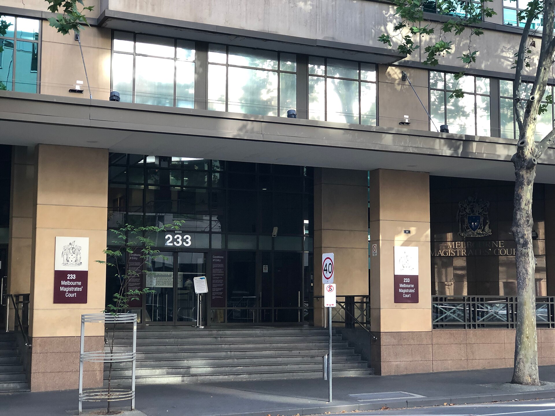 The entrance to the Melbourne Magistrates' Court at 233 William Street: glass doors at the top of wide stone steps between sandstone-clad pillars, with maroon signs and the Victorian coat of arms.