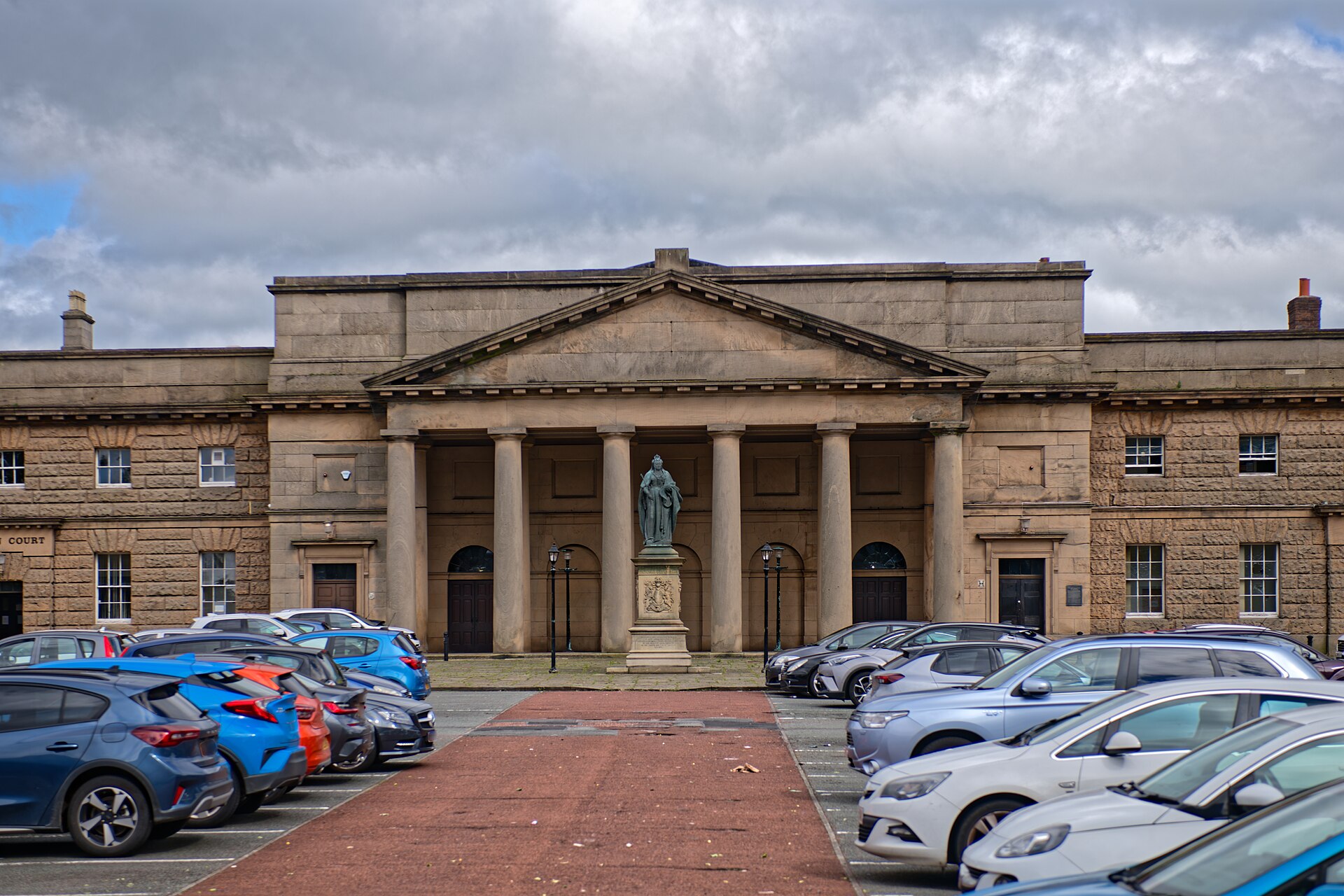 The columned stone portico of the Assize Courts block at Chester Castle, with a bronze statue of Queen Victoria in front and cars parked across Castle Square, under a cloudy sky.