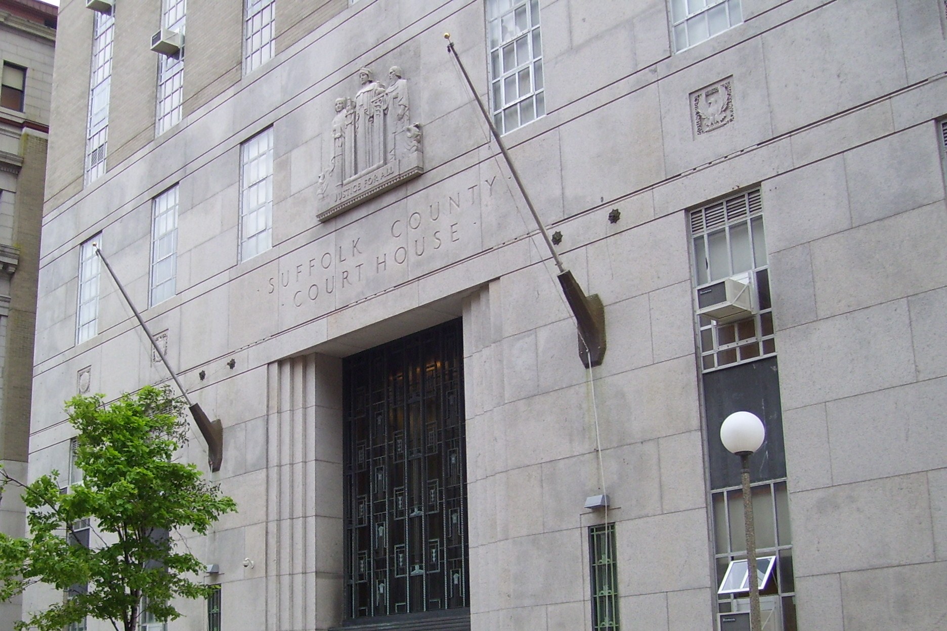 The granite Art Deco front of the Suffolk County Courthouse in Boston, its name carved above a tall latticed entrance beneath a relief panel inscribed 'Justice for All', with two angled flagpoles and a young tree.