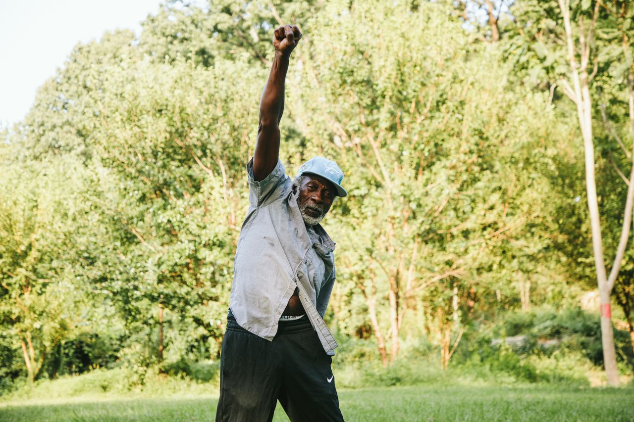 Ervin Harris, an older Black man in a light blue cap and pale shirt, standing on grass before summer trees with his right fist raised.