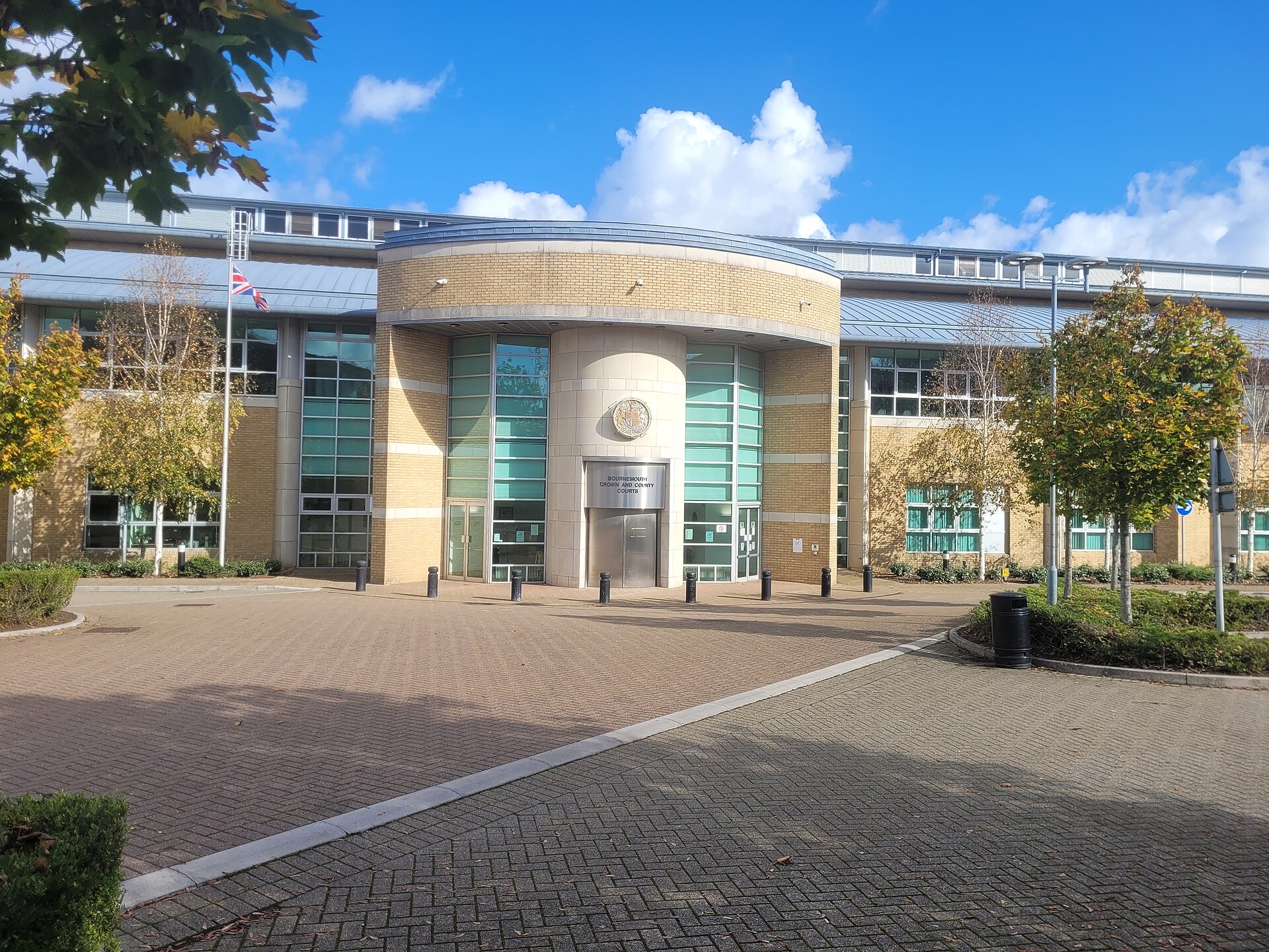 Bournemouth Crown and County Courts on Deansleigh Road: a curved sand-coloured brick entrance with tall green-tinted windows and a royal crest, flanked by autumn trees and a Union flag, under a blue sky.