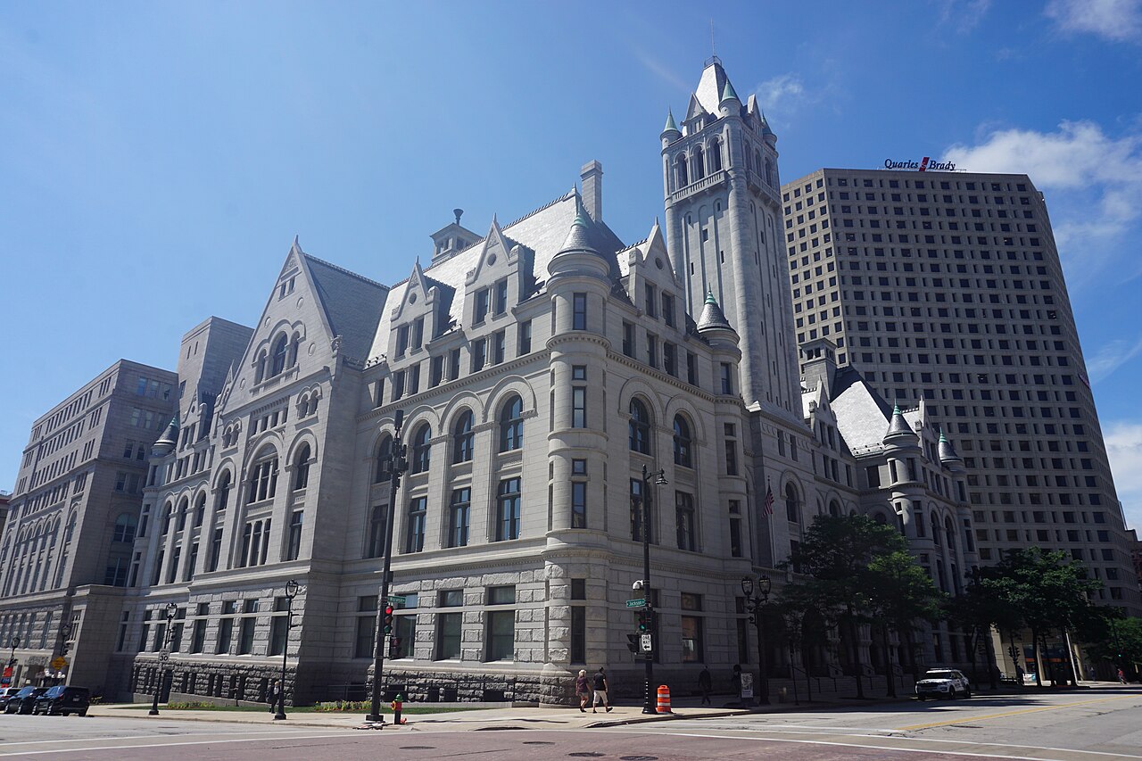 The Milwaukee Federal Building and U.S. Courthouse, a pale granite Romanesque building with steep gables and a tall corner tower, seen from a street corner under a clear blue sky.