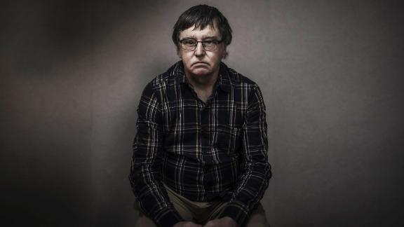 Alan Hall, a man in glasses and a dark checked shirt, sitting against a plain grey wall and looking straight at the camera.