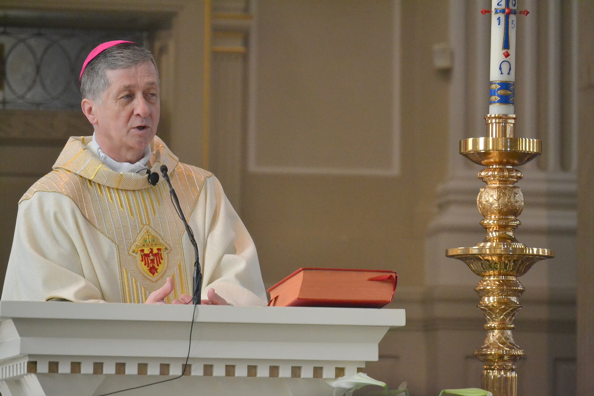 Blase Cupich in cream and gold vestments and a pink zucchetto speaks at a white lectern beside a red book, with a tall gilded candlestick and Easter candle to his left.