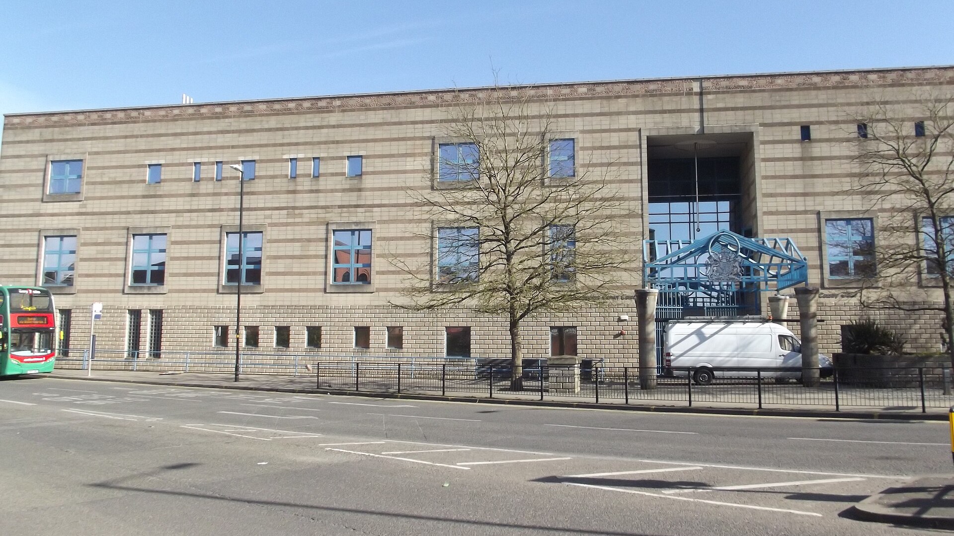 The long, banded sandstone-coloured frontage of Wolverhampton Crown Court on Piper’s Row, with a blue metal canopy and royal coat of arms over the entrance, a bare tree and railings in front, and a green-and-red bus at the left.