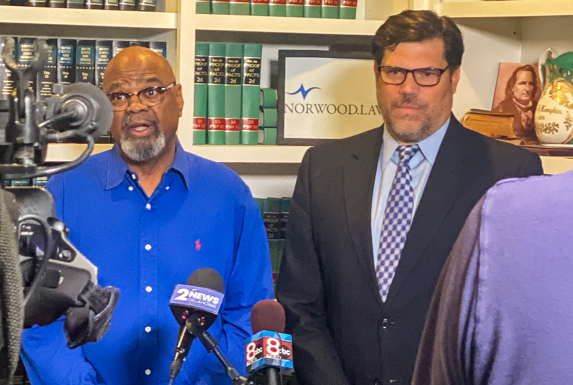 Glynn Simmons, in glasses and a bright blue shirt, speaks into TV station microphones beside his attorney Joe Norwood, in a suit and tie, in front of law-book shelves and a Norwood Law sign.
