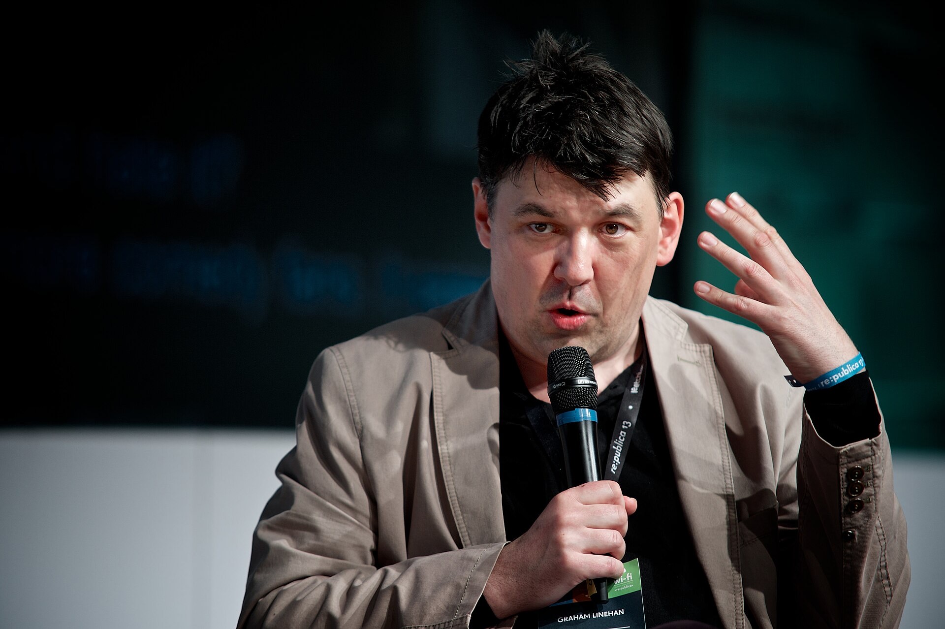 Graham Linehan, dark-haired and wearing a beige jacket over a black shirt and a conference lanyard, speaking into a microphone and gesturing with his left hand against a dark background.