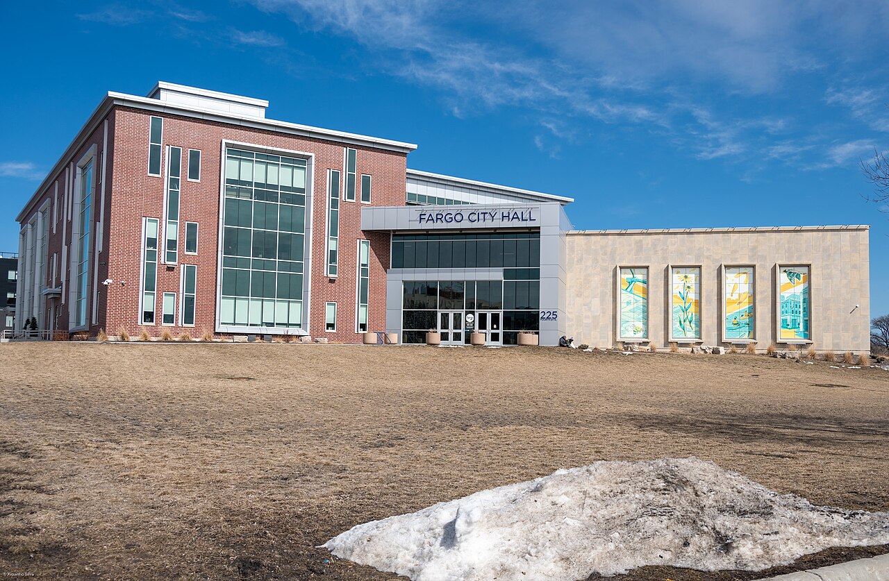 Fargo City Hall in North Dakota on a clear winter day: a two-storey red brick and glass building with a canopy reading “FARGO CITY HALL”, adjoined by a pale stone wing set with four tall panels of stylised prairie artwork, above a dormant lawn with a patch of melting snow.