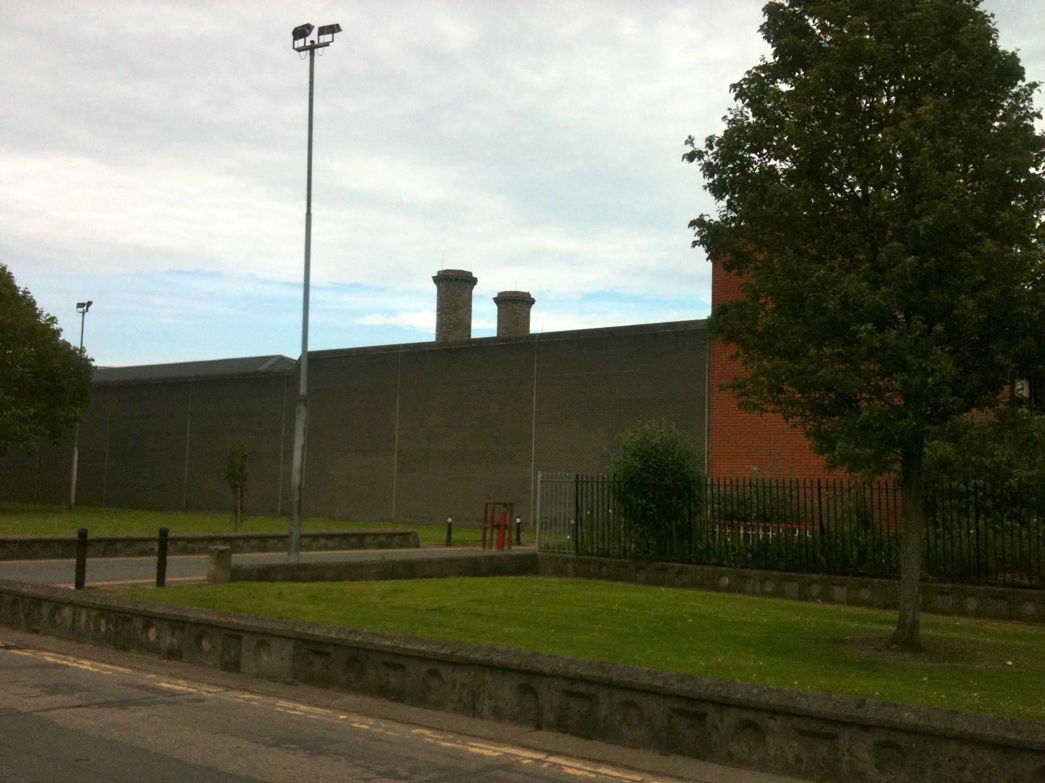 The high grey perimeter wall of Mountjoy Prison in Dublin, with a red-brick building and chimneys behind it, a mown verge, a tall floodlight and a tree in the foreground.