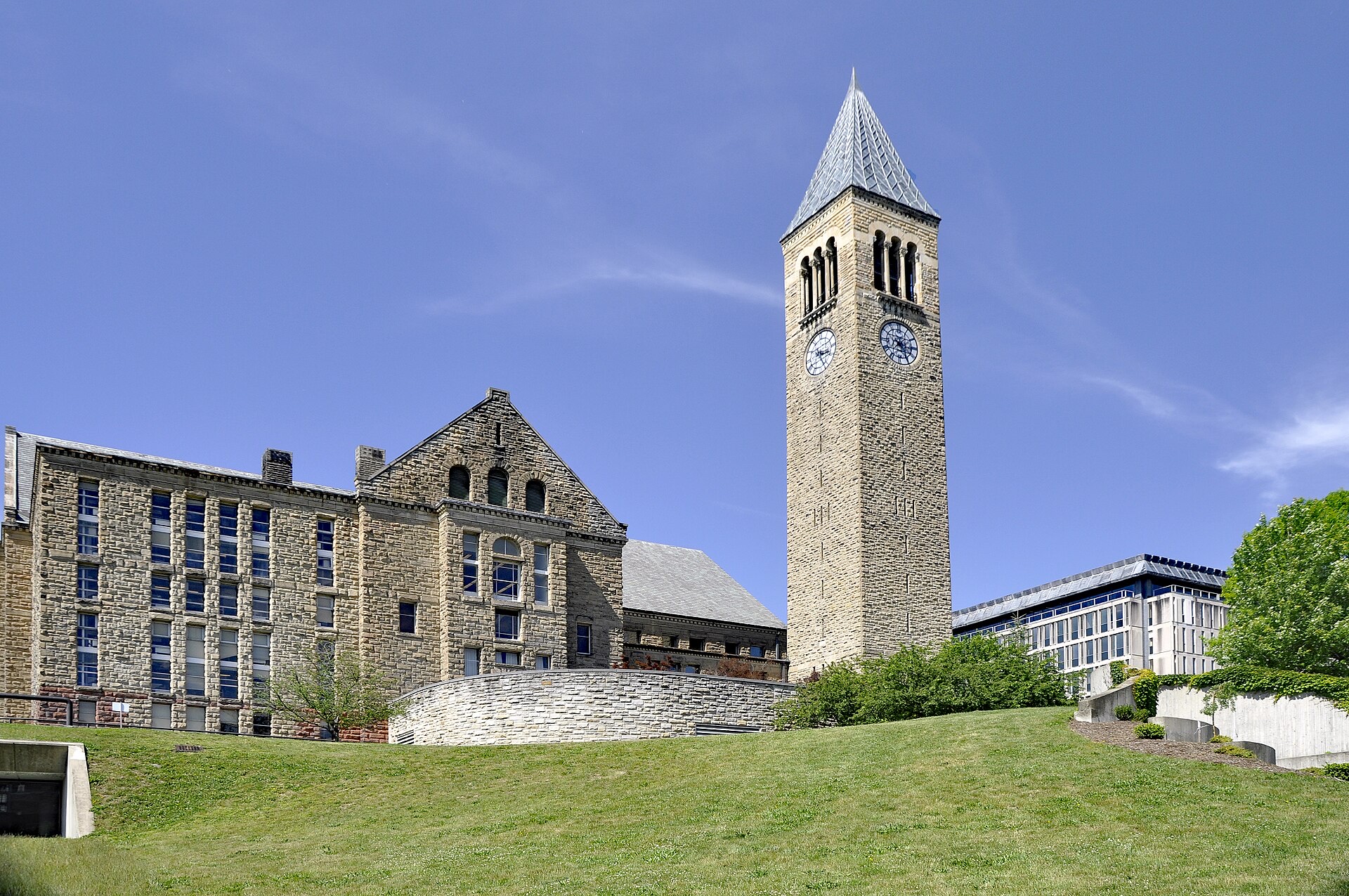 McGraw Tower, a tall stone clock tower with a pointed slate spire, rising between the rough-stone Uris Library and the modern Olin Library on Cornell University's campus, above a grassy slope under a blue sky.