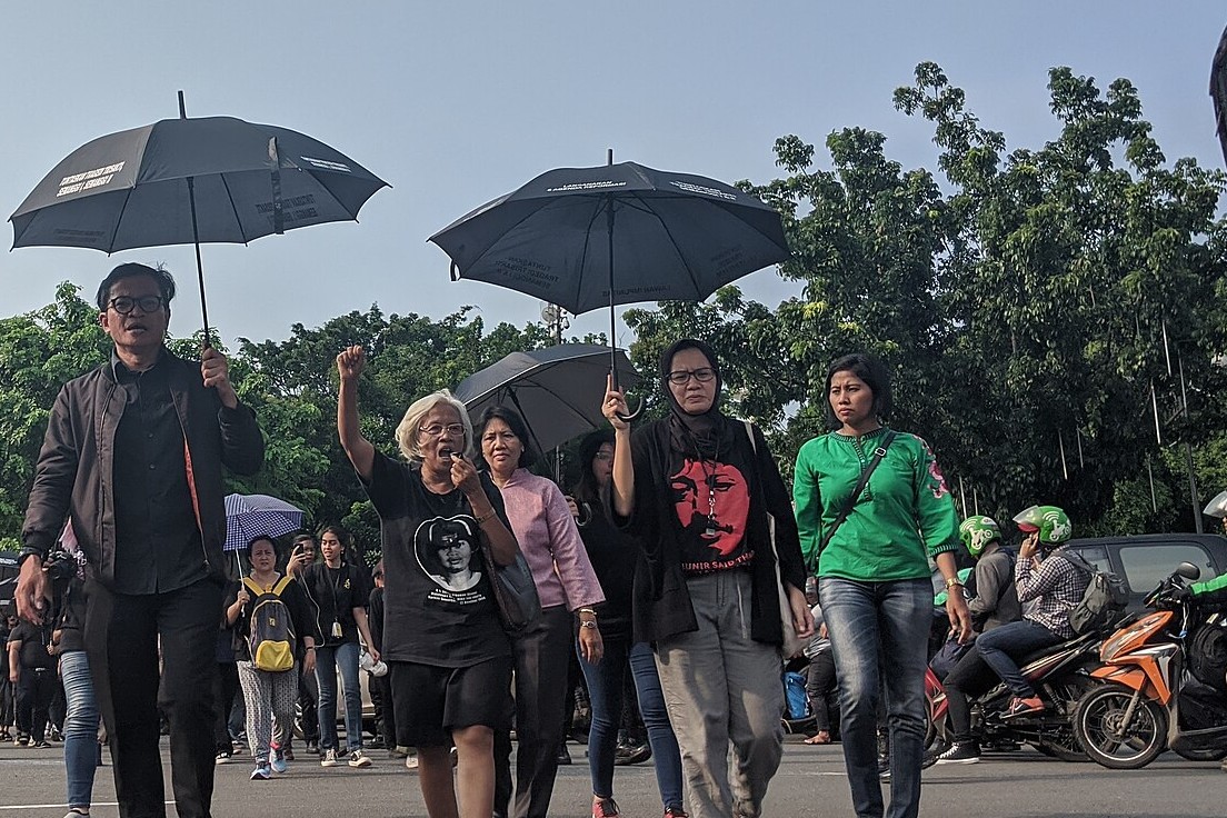 Usman Hamid, in black and holding a black umbrella, walks across a Jakarta road beside Maria Catarina Sumarsih, who raises a fist and speaks into a megaphone, with other women in black and a marcher in a Munir T-shirt, trees and motorcycles behind.