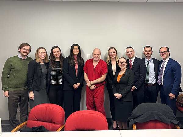 Jason Hubbell, in a red prison uniform and handcuffs, stands in the middle of a row of ten Notre Dame Exoneration Justice Clinic staff and students in business attire, in a plain room with red chairs in the foreground.