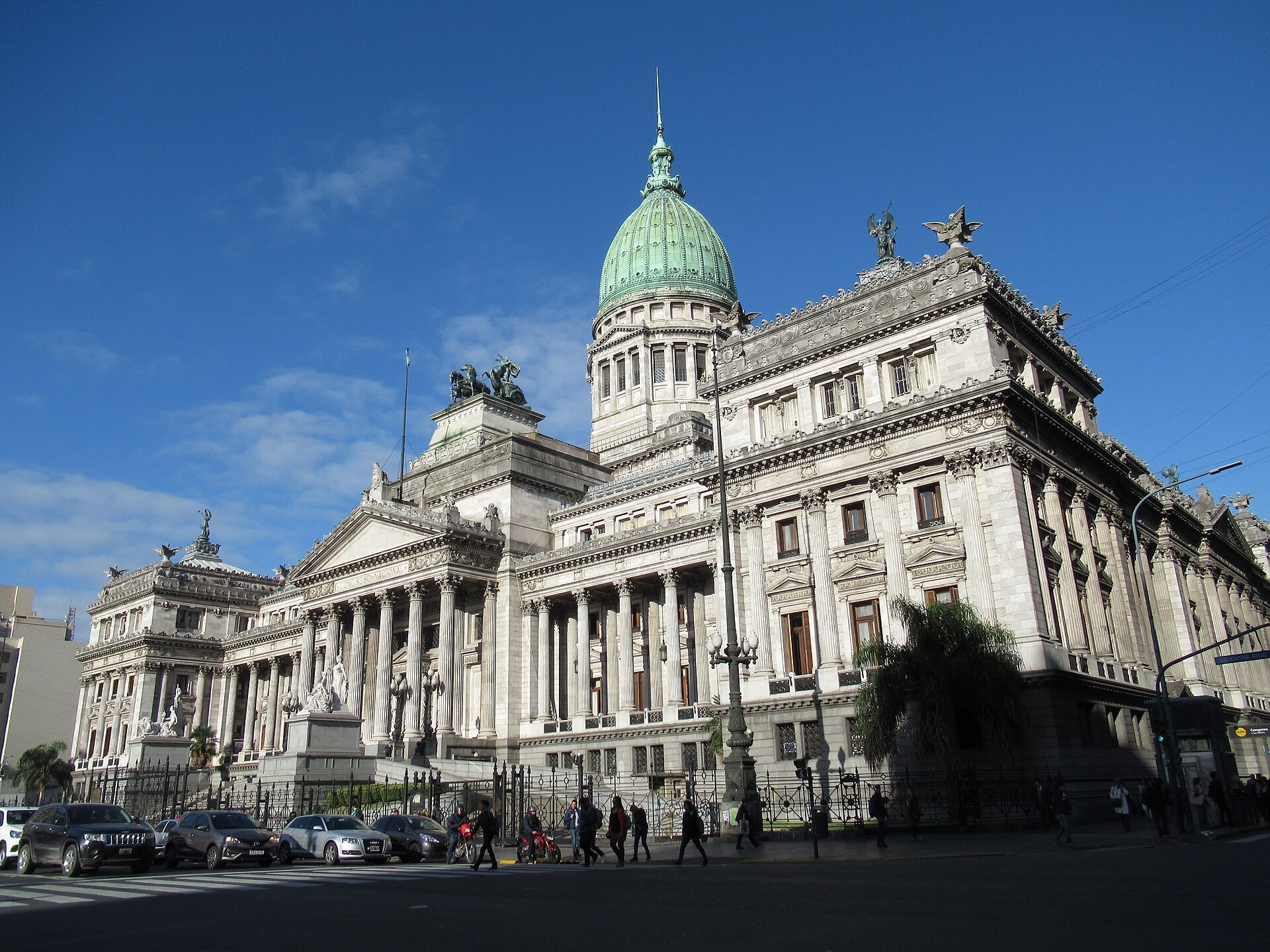 The Palacio del Congreso in Buenos Aires, seat of the Argentine national legislature, its pale stone facade and colonnaded portico rising to a green copper dome against a clear blue sky, with pedestrians passing on the pavement in front.