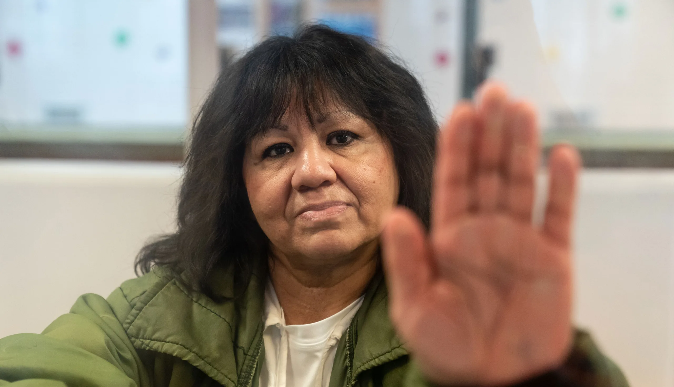 Melissa Lucio, a woman with shoulder-length dark hair in a green jacket over a white shirt, looks into the camera while holding up her open palm toward the lens.