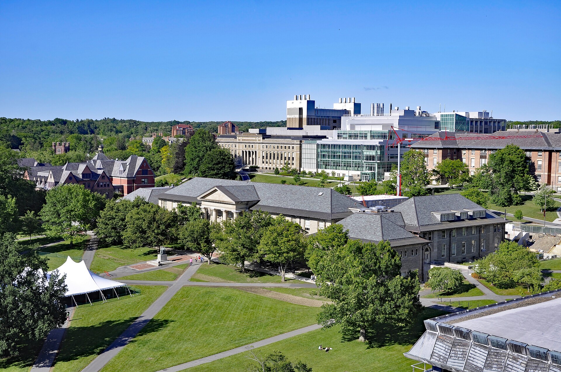 An elevated view across Cornell University's Arts Quad on a sunny day: lawns and paths lined with trees, Goldwin Smith Hall with its columned portico, and newer science buildings beyond.