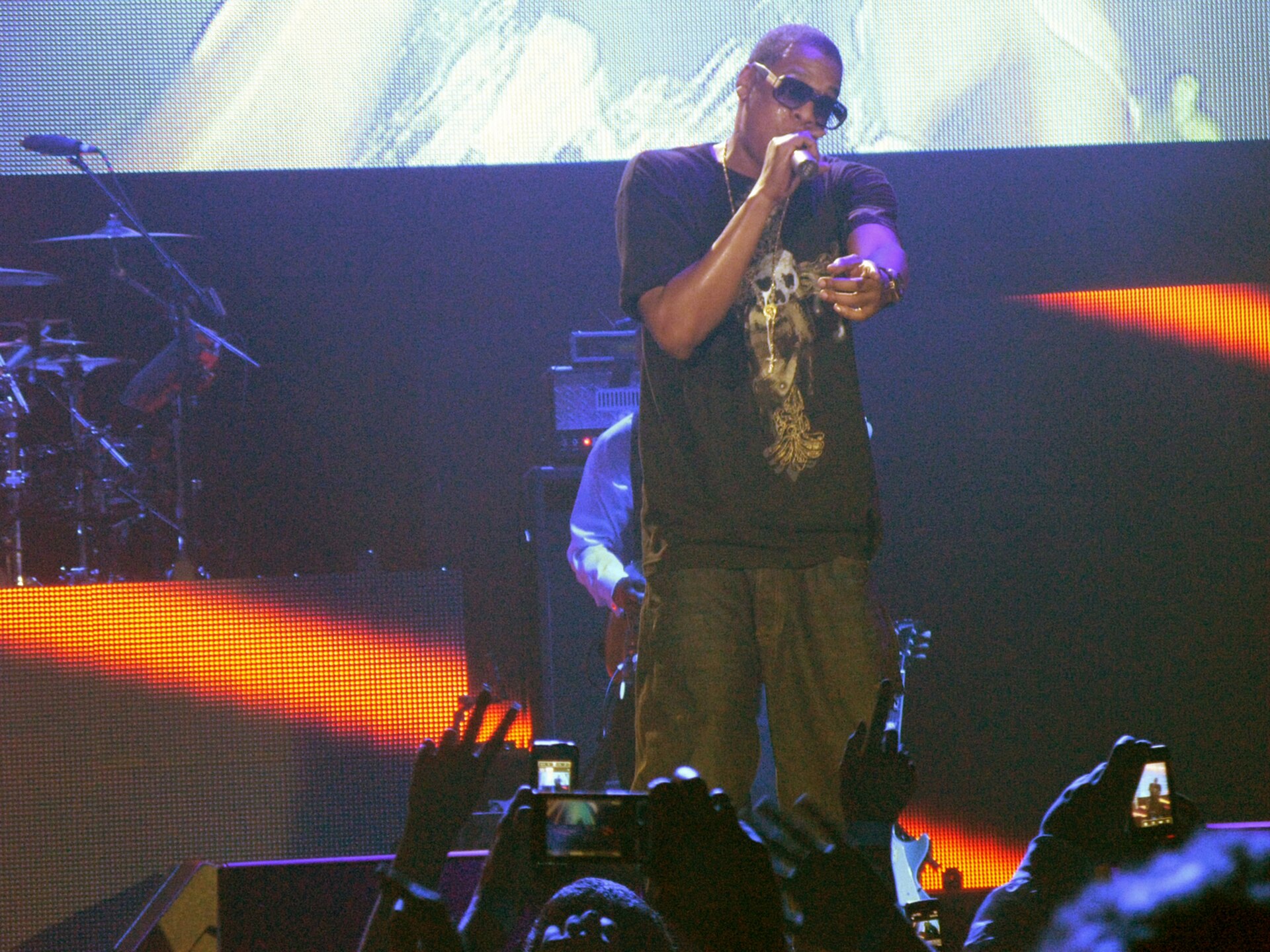 Jay-Z performing on stage in sunglasses and a dark T-shirt, holding a microphone and pointing towards the crowd, with fans' raised hands and phone screens in the foreground.