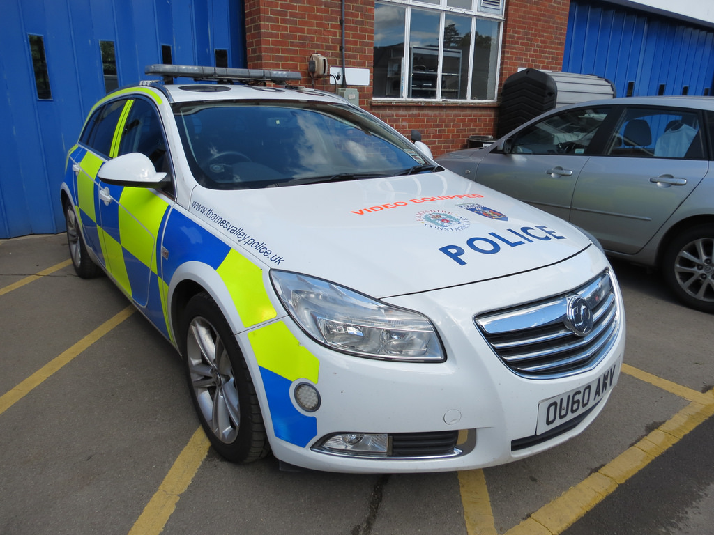 A Thames Valley Police estate car in yellow-and-blue Battenburg livery, with POLICE and the force's web address on the bonnet, parked in a yard beside a brick building.