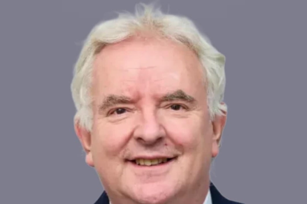 Head-and-shoulders portrait of Richard Clayton KC, a smiling man with swept-back white hair in a dark suit and striped tie, against a plain grey background.