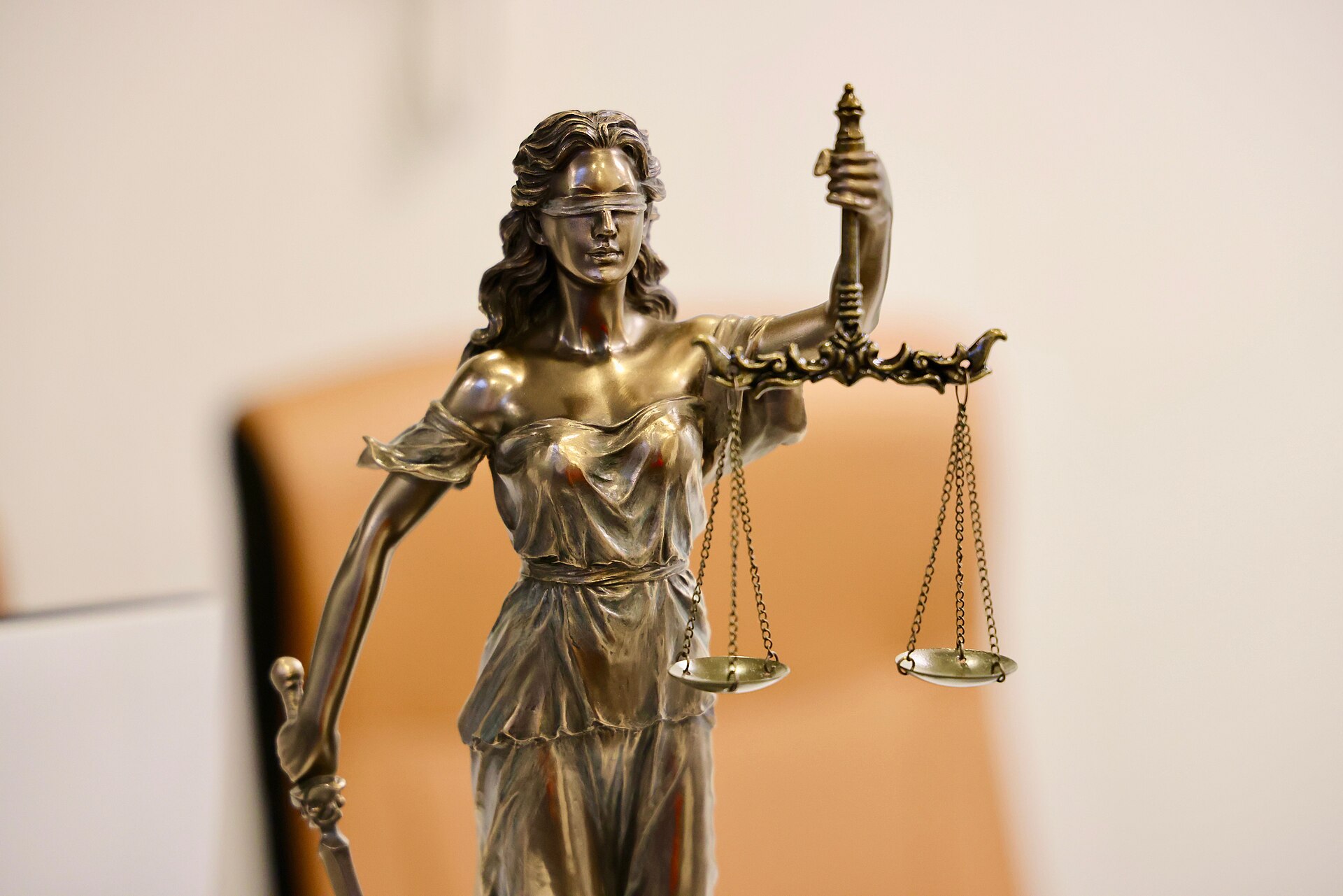 A bronze statuette of Lady Justice, blindfolded, holding a sword in one hand and balanced scales raised in the other, against a soft blurred background.