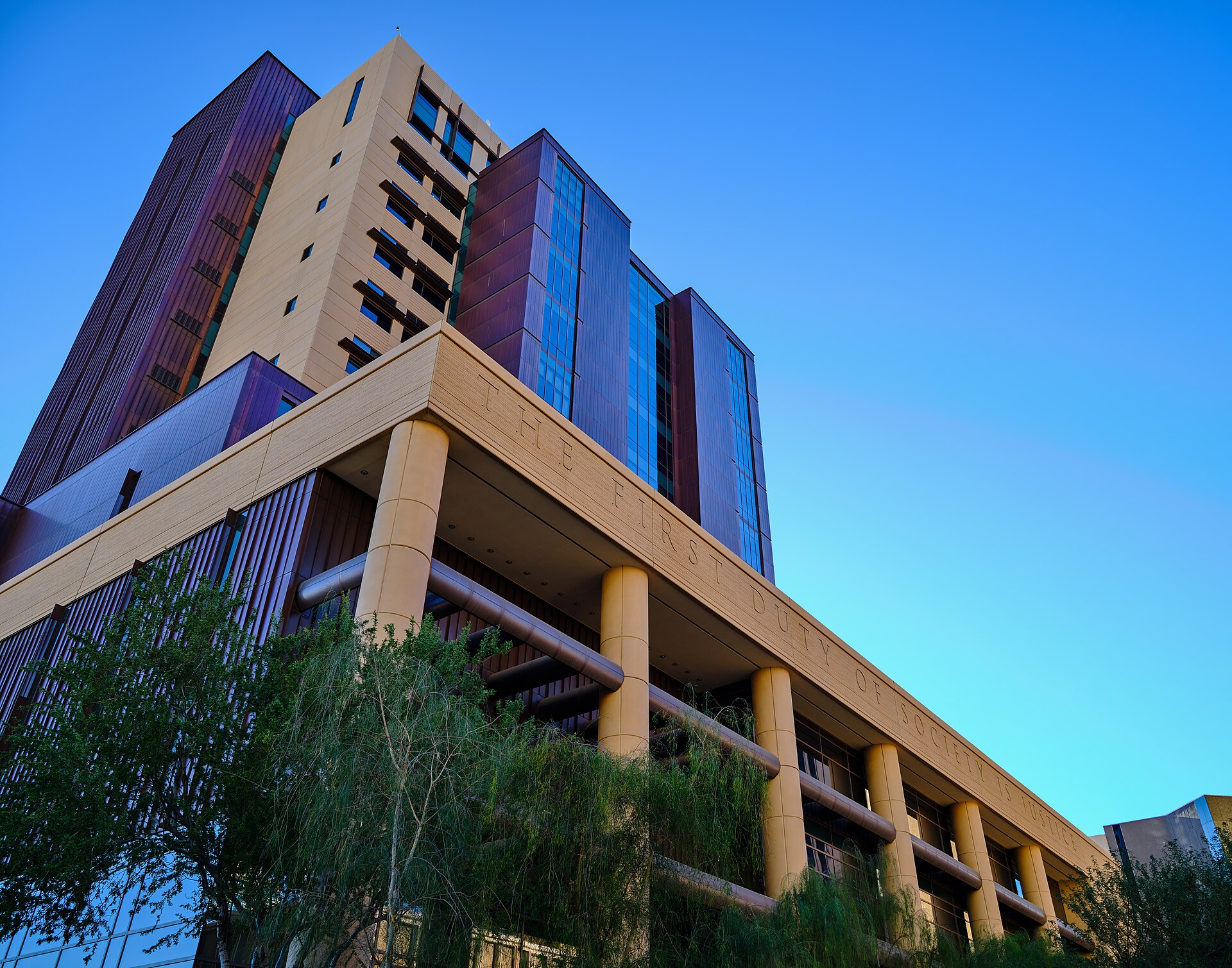 Looking up at a Superior Court of Maricopa County tower in downtown Phoenix, its long sandstone-coloured frieze carved with THE FIRST DUTY OF SOCIETY IS JUSTICE above round columns, under a clear blue sky.