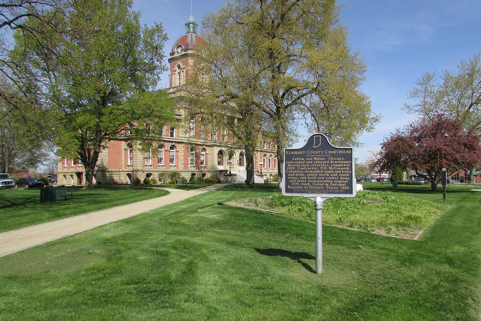 The historic red-brick Elkhart County Courthouse in Goshen, Indiana, with a domed clock tower, behind spring trees and a lawn, with an Indiana historical marker in the foreground.