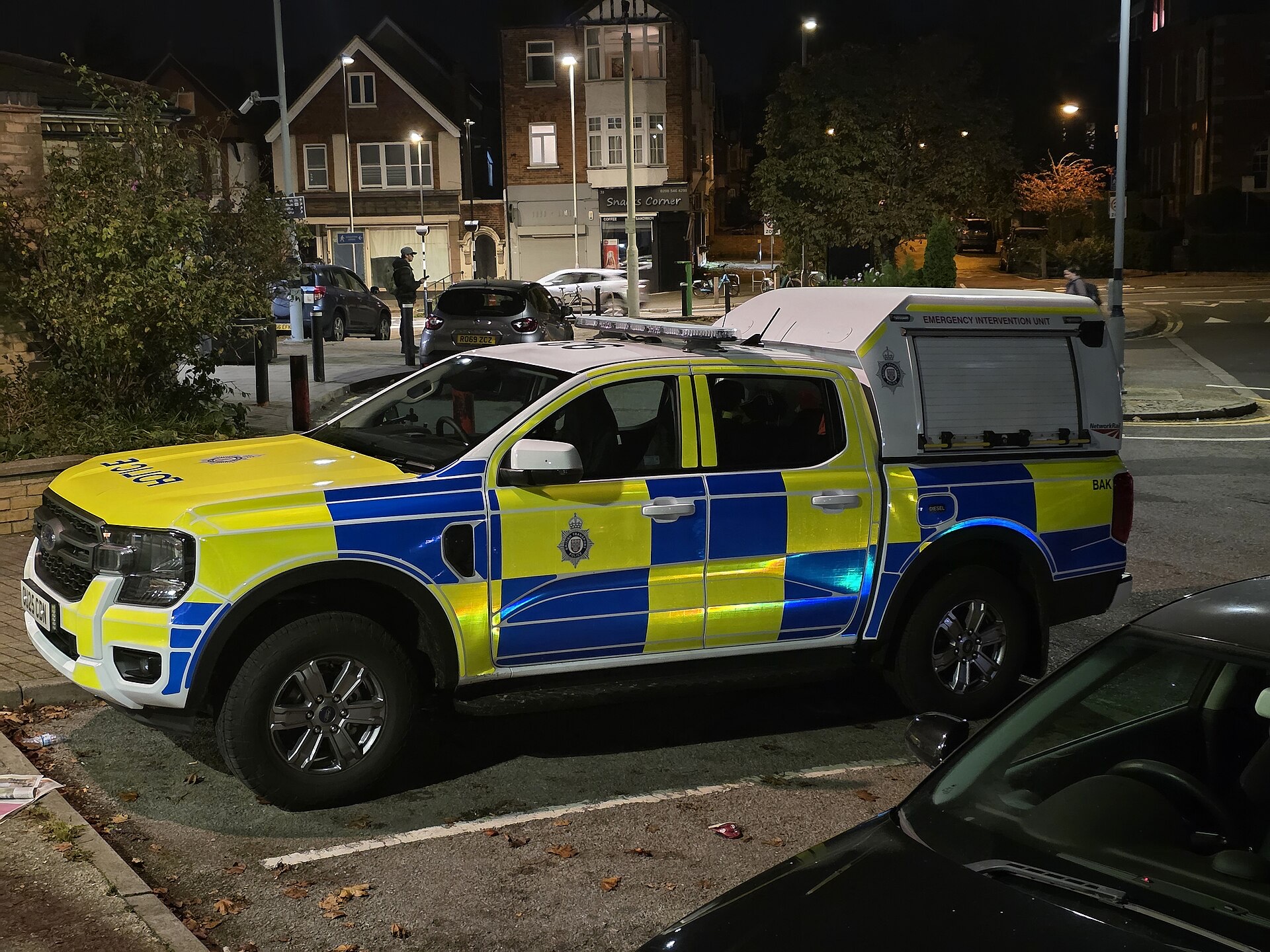 A British Transport Police Emergency Intervention Unit pick-up in yellow-and-blue Battenburg livery, with the force crest on its door, parked by a station at night.