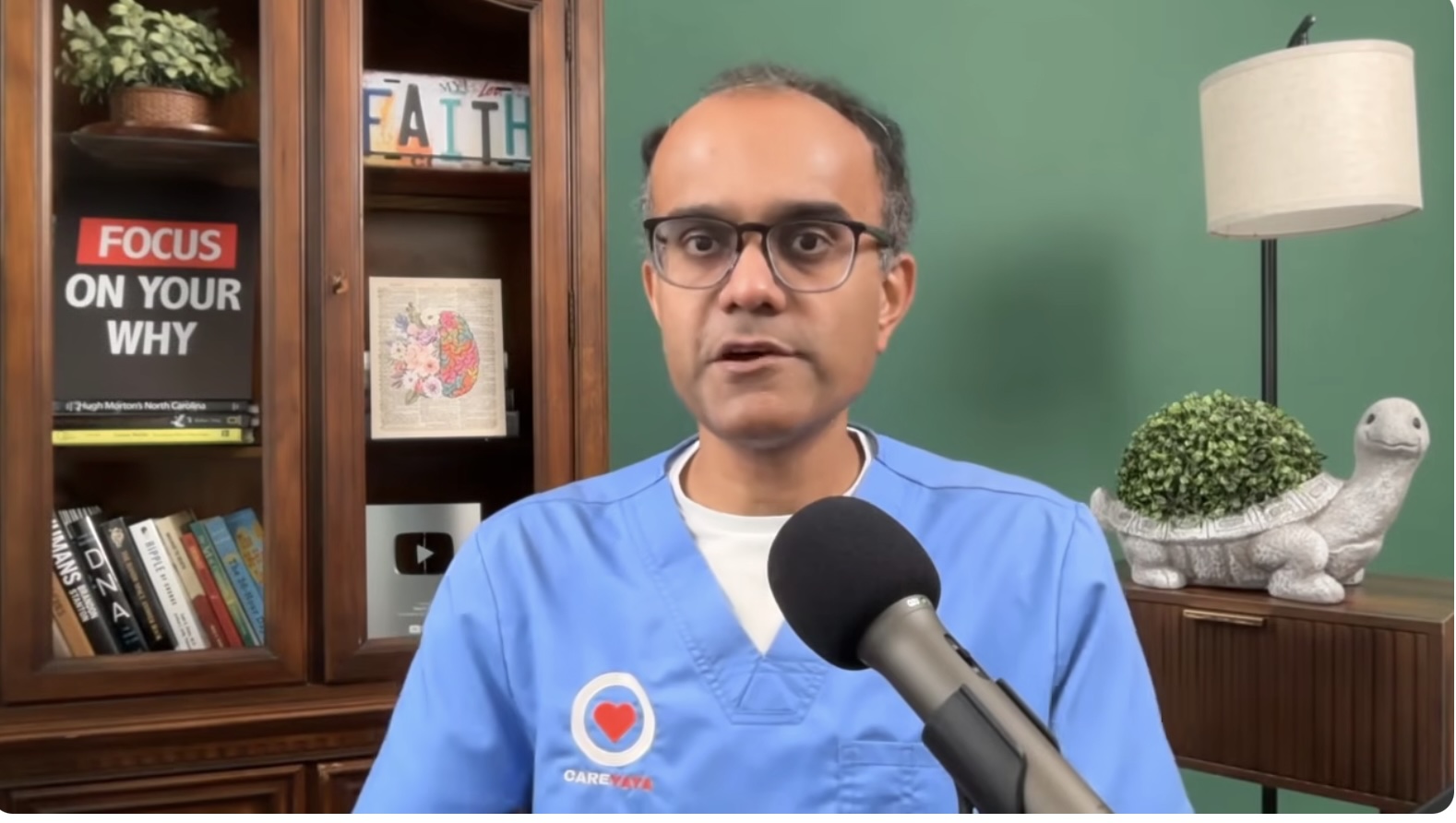 Neal K. Shah, a bespectacled man in blue CareYaya-branded medical scrubs, speaking to a podcast microphone in front of a bookcase and a green wall.